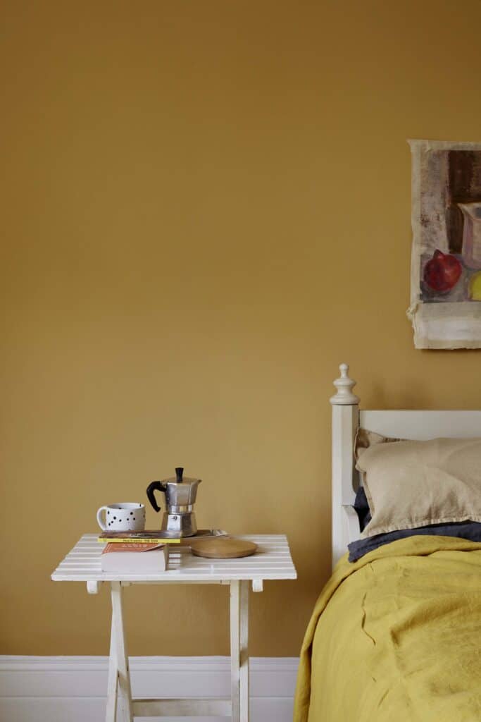Yellow bedroom wall with white bedside table with coffee pot and cup in front. You can also see the corner of a white bed frame and yellow duvet.