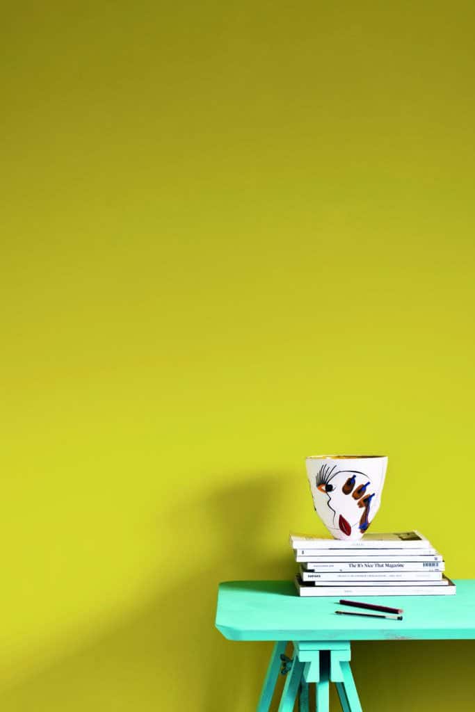 A bright yellow painted wall with a turquoise table in front. On top of the table is a mug and some books.