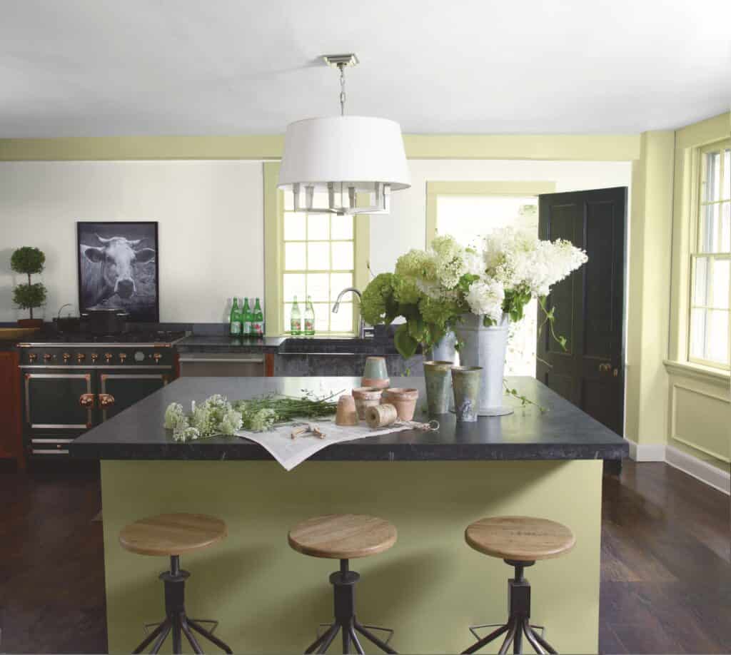 Large kitchen island with three bar stool stood in front. The island is painted sage green. There is a vase of flowers on top of the island and a kitchen in the background