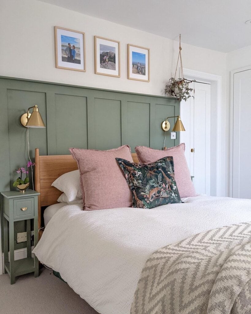 A green panelled bedroom wall that acts as an oversized headboard behind a wooden bed that is dressed with pink cushions and a chevron blanket. THere are conical lamps either side and three small pictures above the bed.