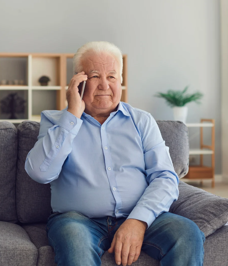 Older man in blue shirt holding phone sits on grey sofa