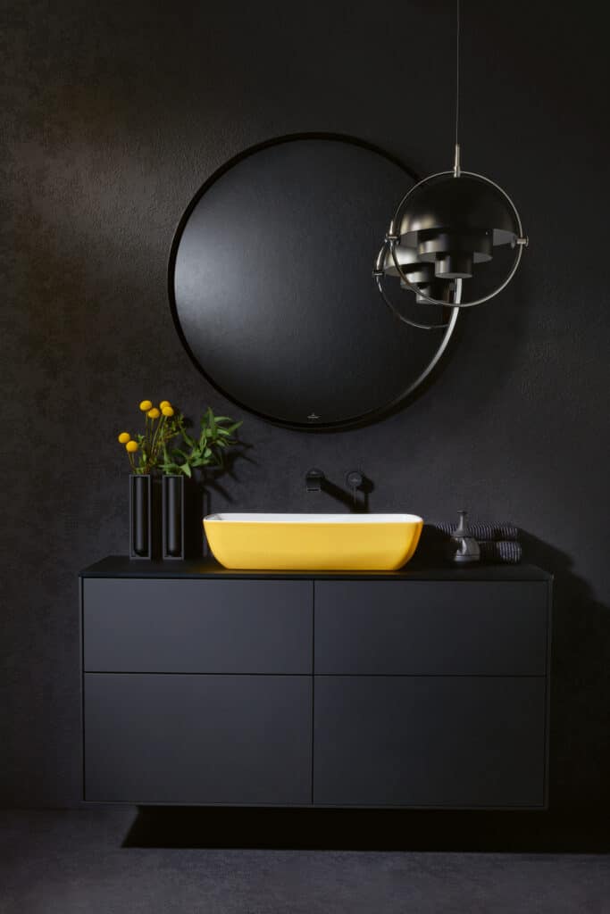 Black wall with round birror, black bathroom unit and yellow sink basin on top. Vase of flowers sat next to sink. modern