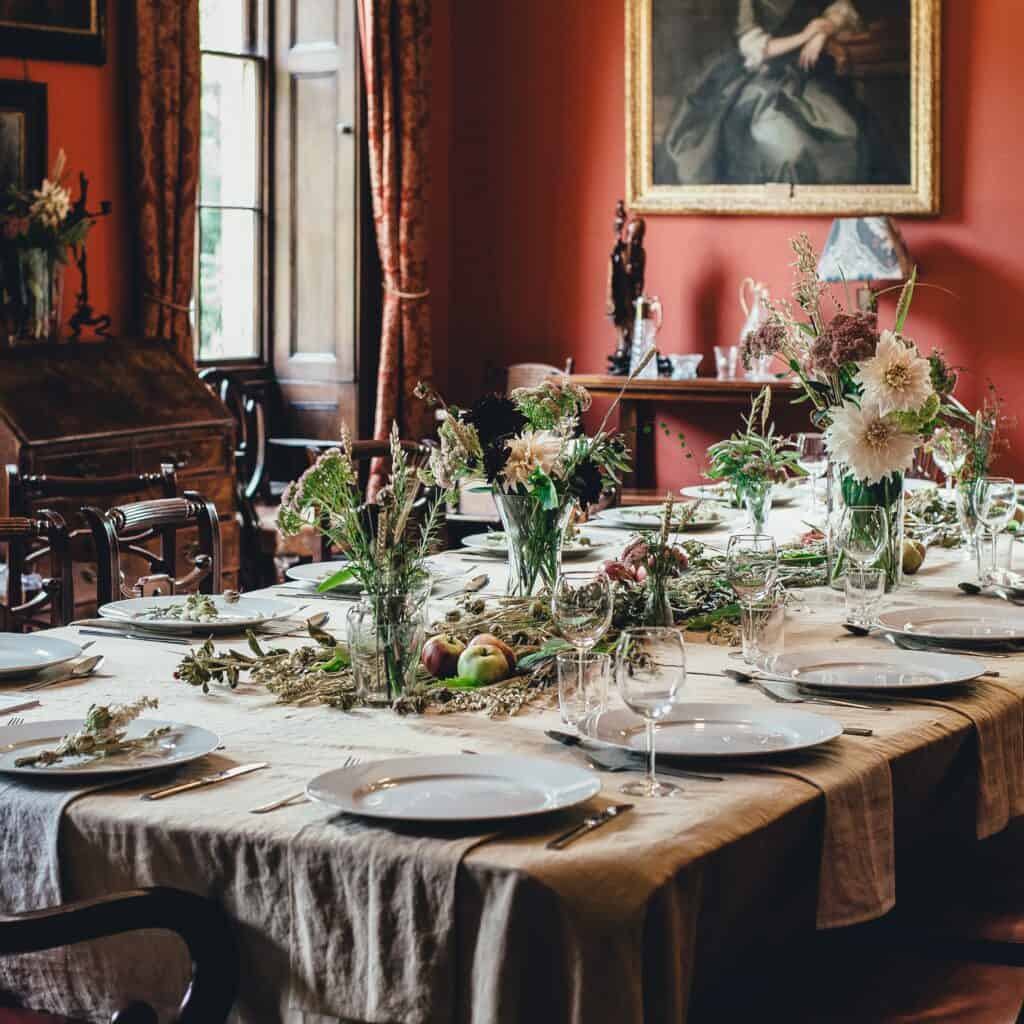 A fully laid table with vases of floiage, plates and a cream tablecloth. The room is painted dark red and is a traditional setting.