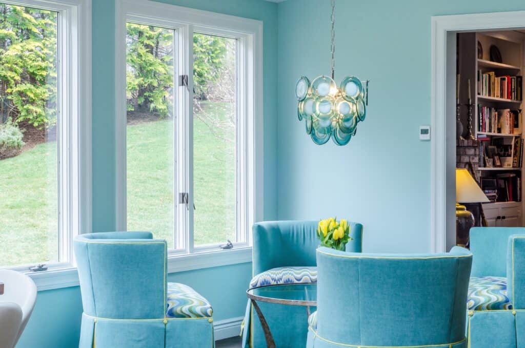 Blue velvet chairs grouped together. Above them is a glass art decor style light and you can see through to a glimpse of al ibrary