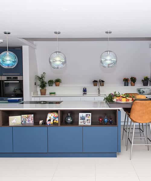 Over size kitchen island with blue fronted cabinets and open dark wood shelving. There are large glass lights above the island and orange leather breakfast stools. In the background are various plants on the sink shelf.