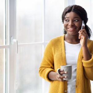 Black lady wearing white tshirt and yellow cardigan on mobile phone. She is holding a cup of tea.