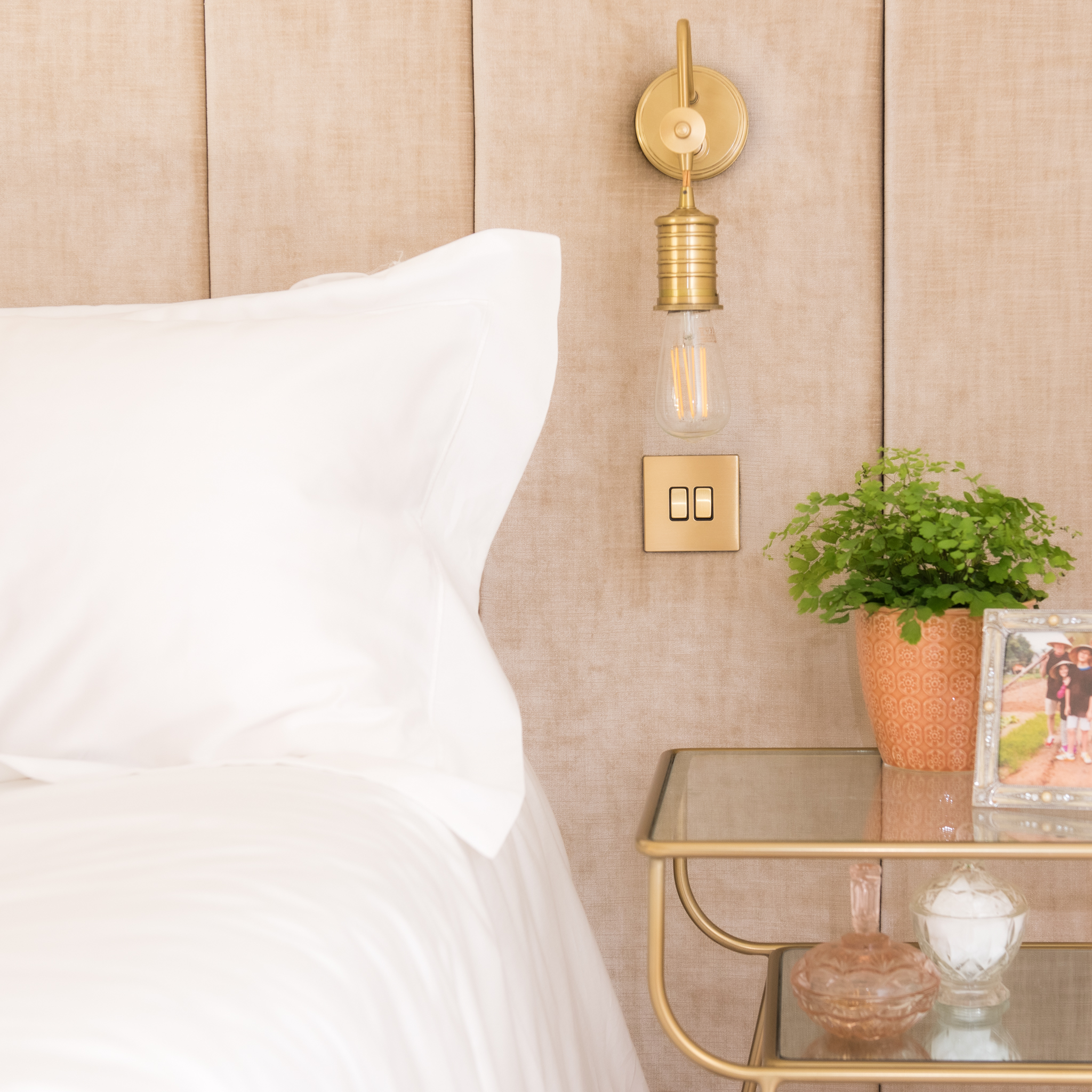 White bed next to gold & glass table and gold lamp on peach headboard
